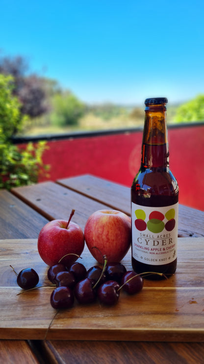 A bread board with cherries, apples and a bottle of Apple and Cherry Non-Alcoholic Ciders