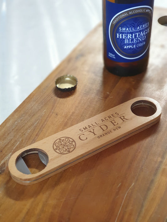 Bamboo Bottle Opener on a wooden table beside a bottle of Small Acres Heritage Blend Apple Cider with a blue label