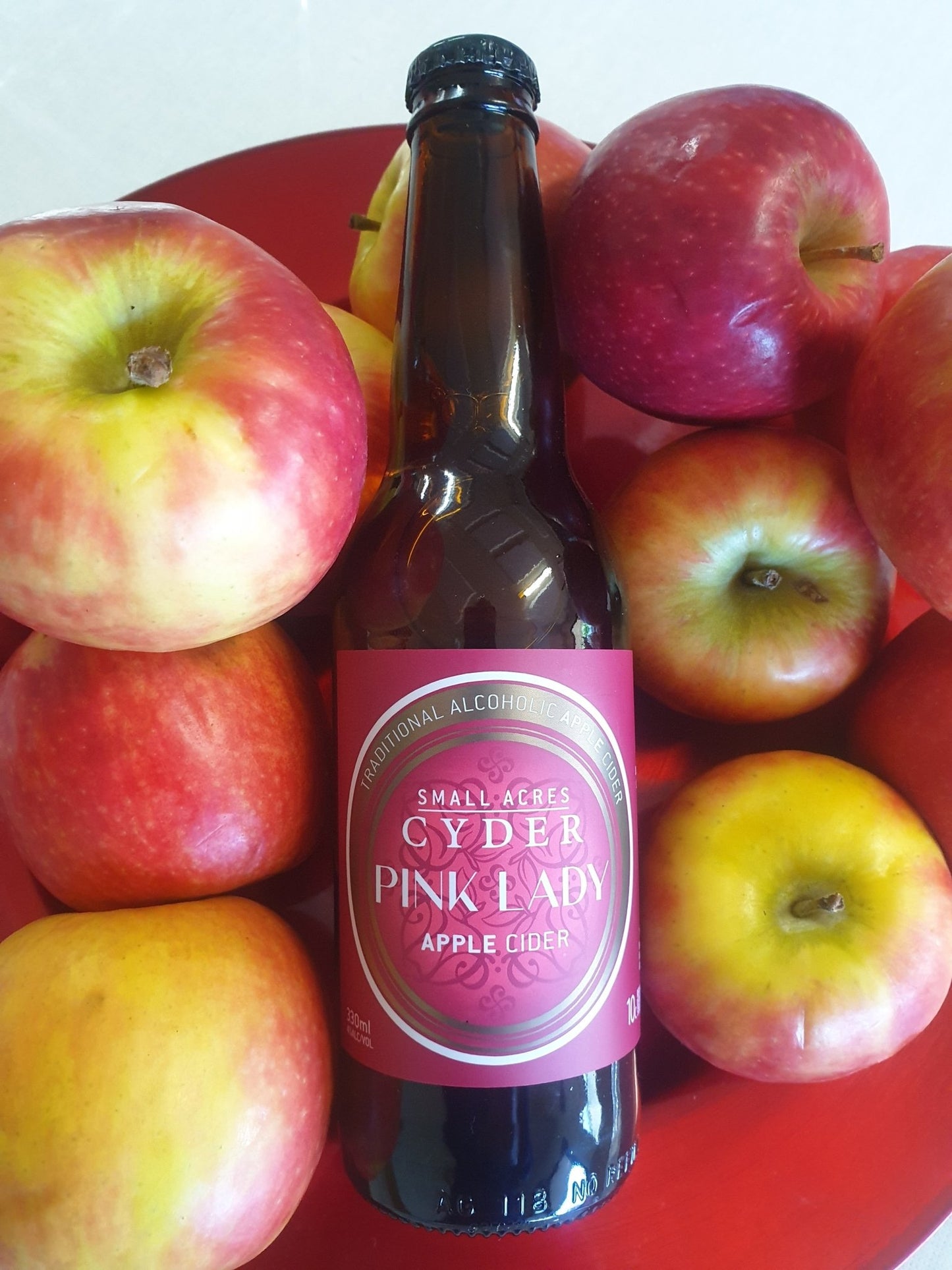 A bottle of Small Acres Cyder Pink Lady on a red plate surrounded by fresh Pink Lady apples