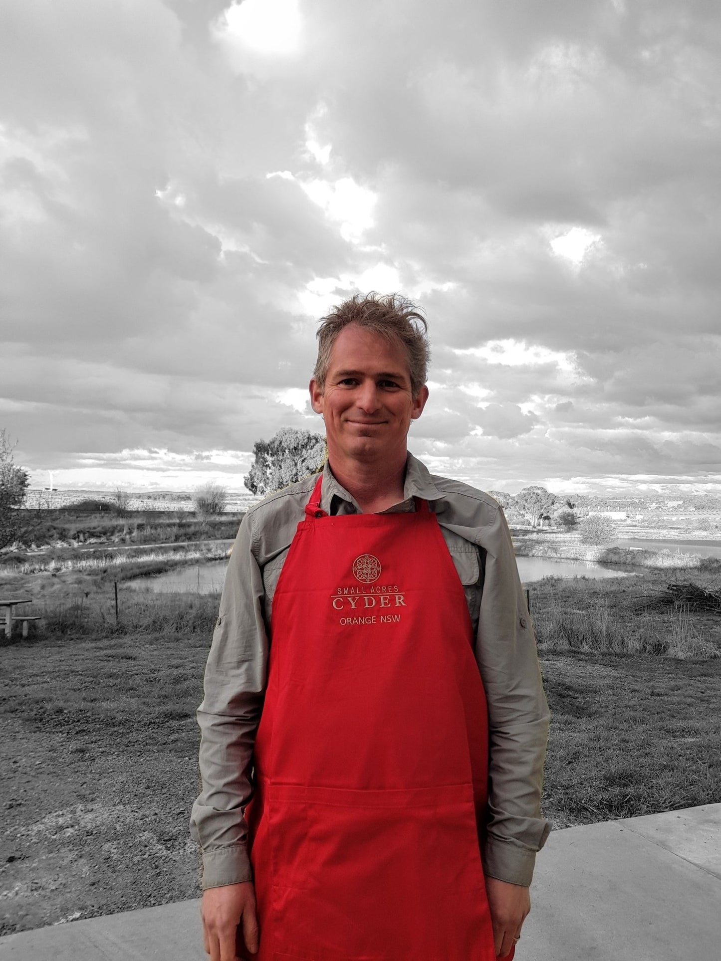 A man with umkempt hair stands before a clouded rural backdrop wearing a red Apron. Most of the image is monochrome except the Red apron and pink face and hands of the model - Small Acres Cyder